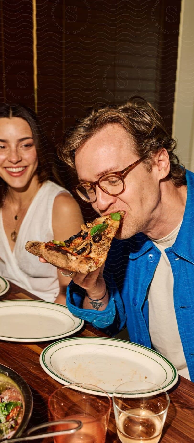 A couple sharing a meal at a restaurant table, the man taking a bite of pizza